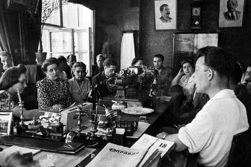 Black and white photo showing people seated around a table with newspaper Pravda in the foreground and portraits of Stalin and Lenin on wall