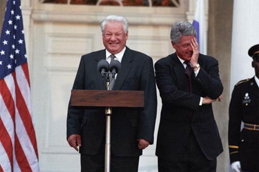 Boris Yeltsin standing at podium while Bill Clinton looks on with hand on his face
