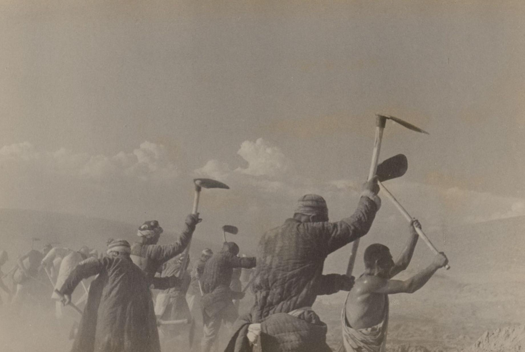 Large group of workers wielding ketmen shovels, surrounded by sky, sand, and mountains in the distance.