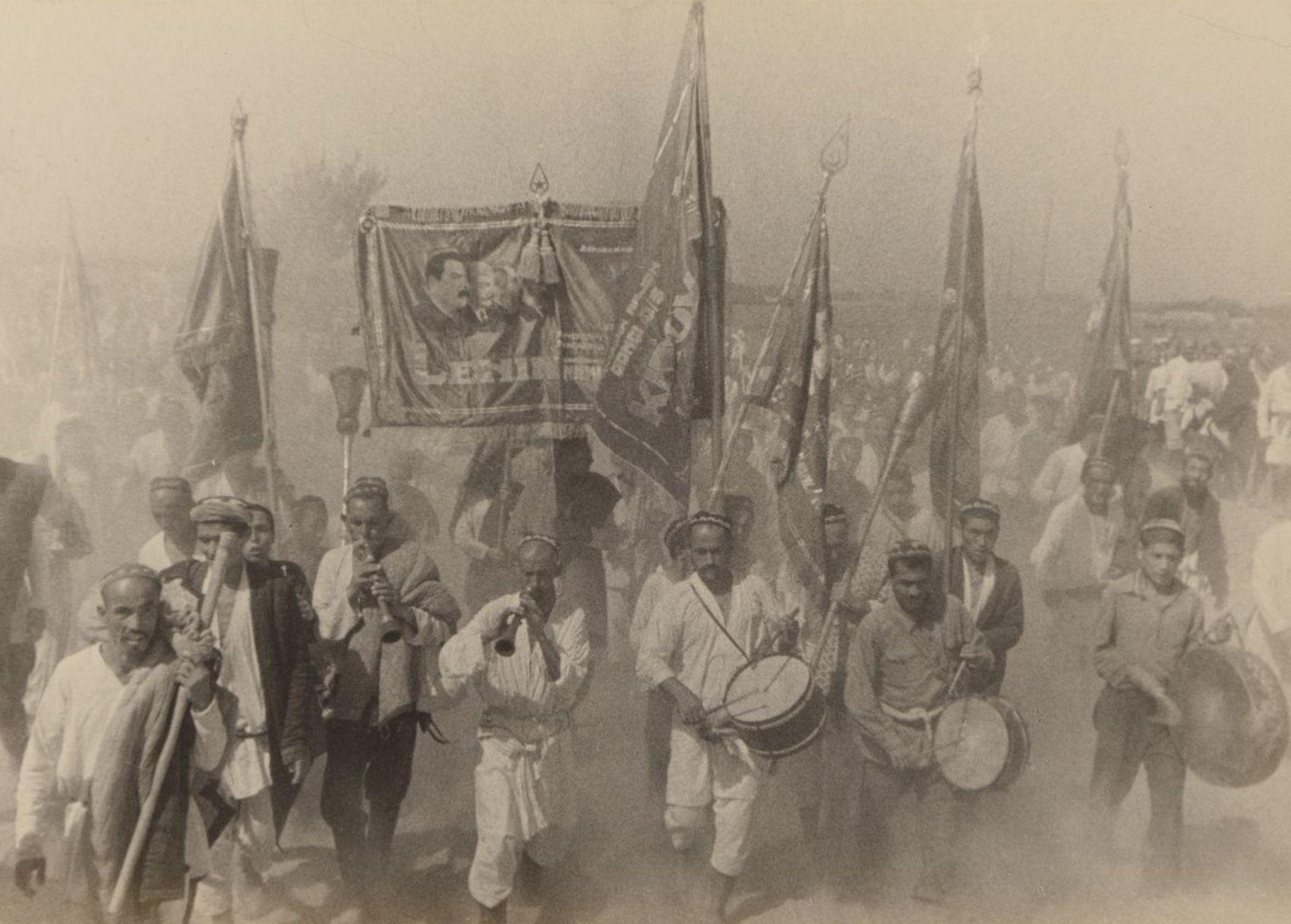 Workers march toward construction site, playing drums and carrying banners.