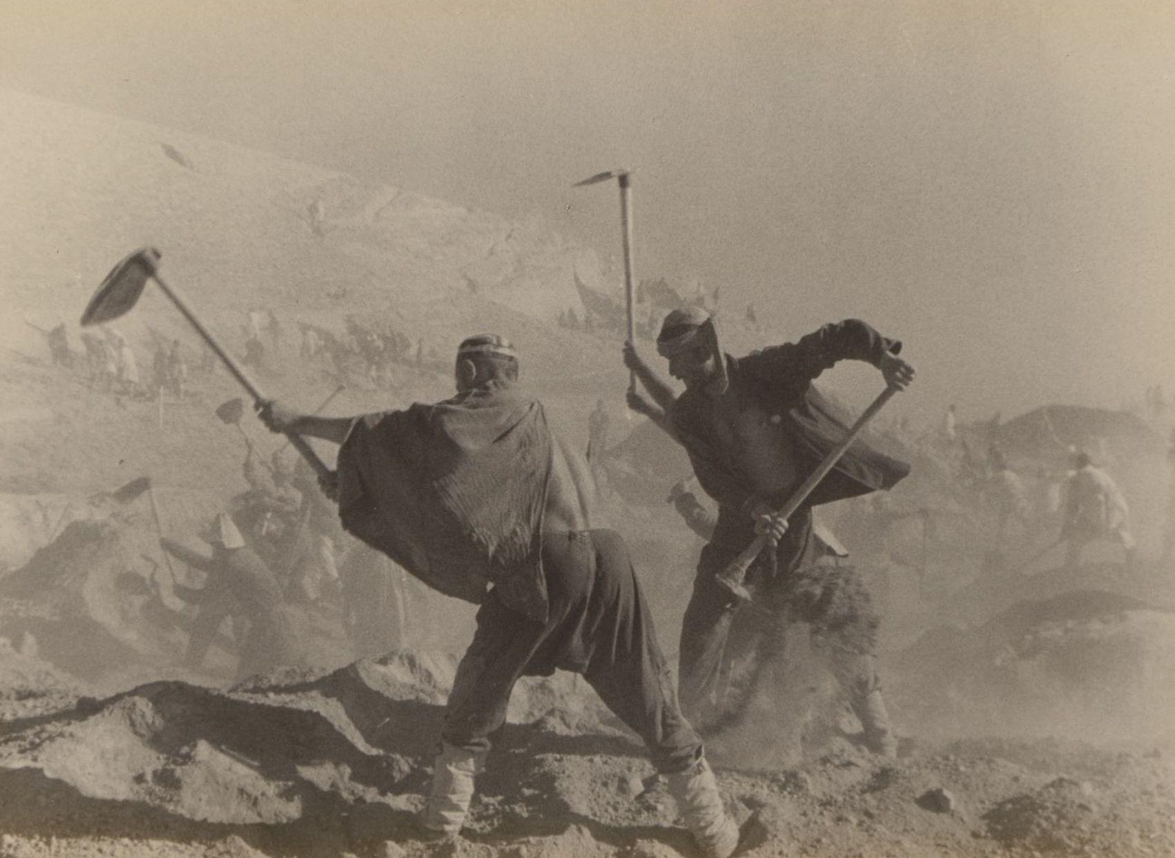 Two workers striking vigorously with their shovels in the foreground; others are visible in the background, partly obscured by the dust.