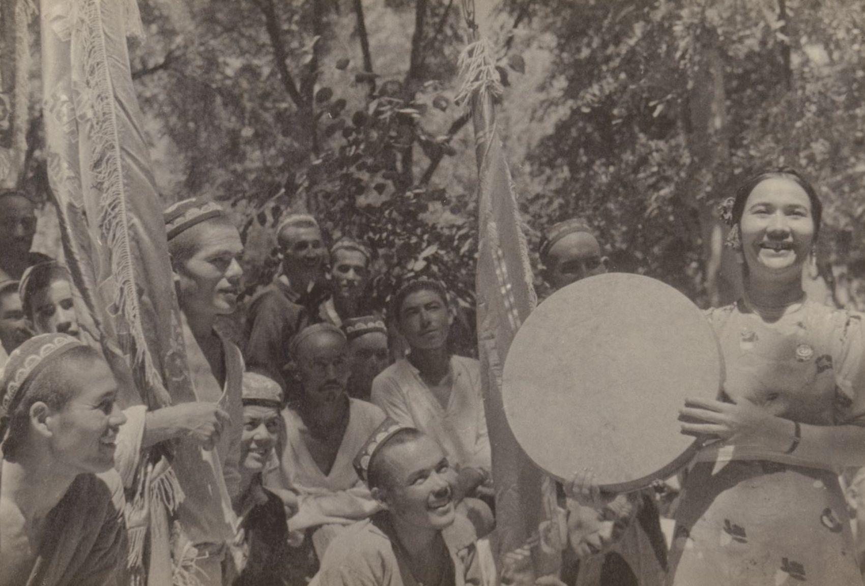 Sarymsakova, smiling, sings and holds drum; a group of smiling workers watch her, some holding banners.