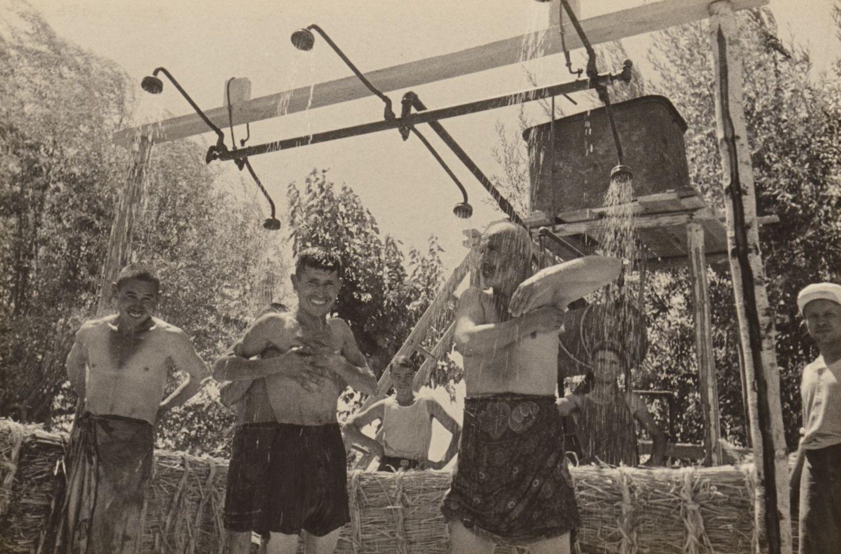 Three smiling workers shower outdoors, wearing underwear or sarongs. Others look on.