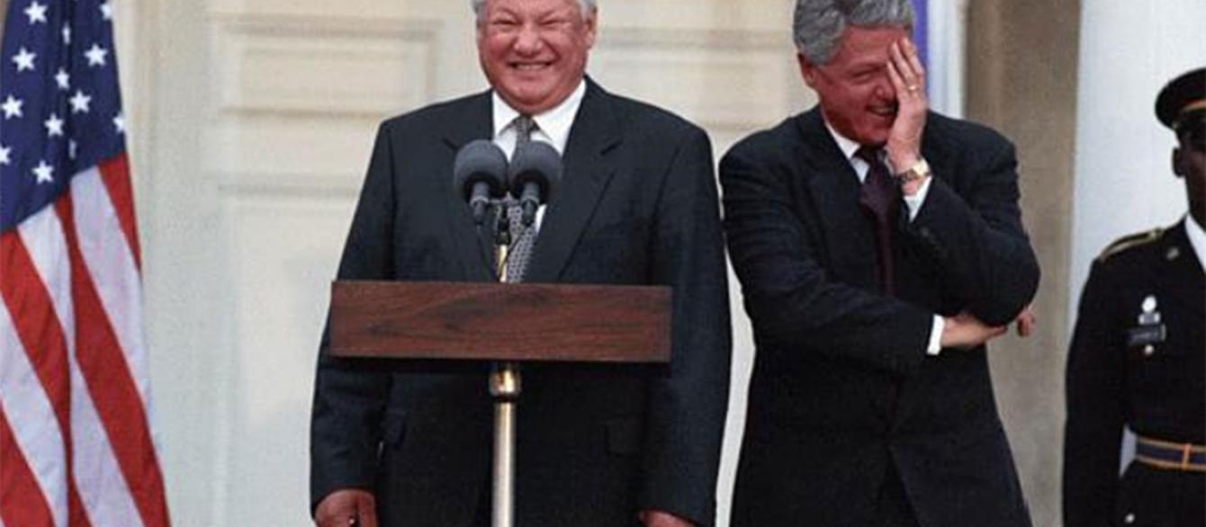 Boris Yeltsin standing at podium while Bill Clinton looks on with hand on his face