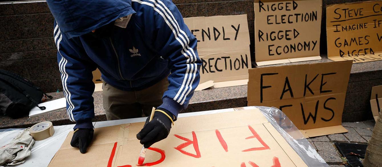 Person in hoodie painting an "America First" sign on cardboard