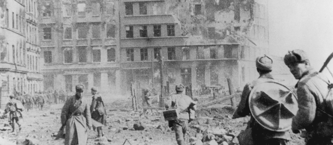 An image of WWII-era Germany, where a building crumbles ahead of a group of soldiers.