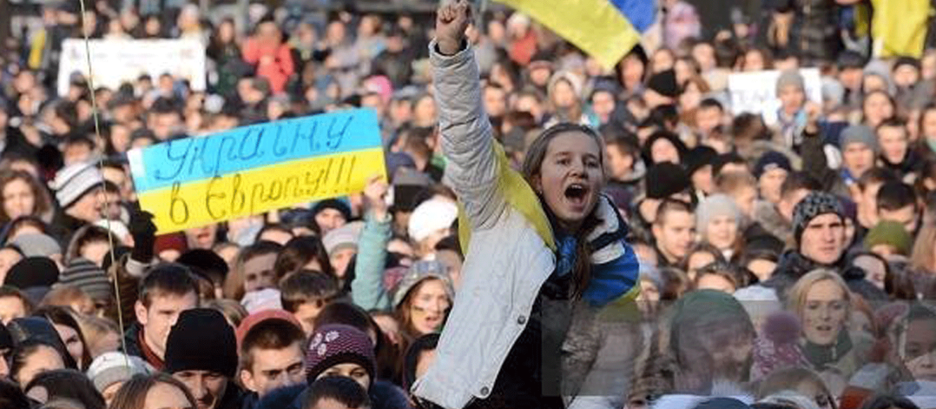 A crowd of people passionately holding Ukranian flags and written signs, centered on a young woman being carried on someone's shoulders, high above the crowd.