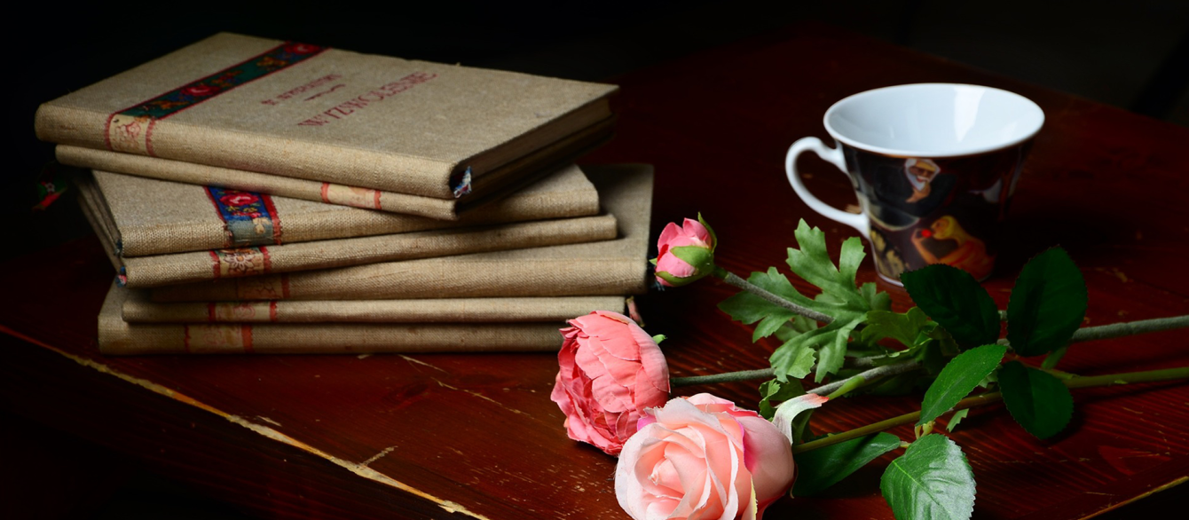 A stack of books next to a small mug and a few pink roses.