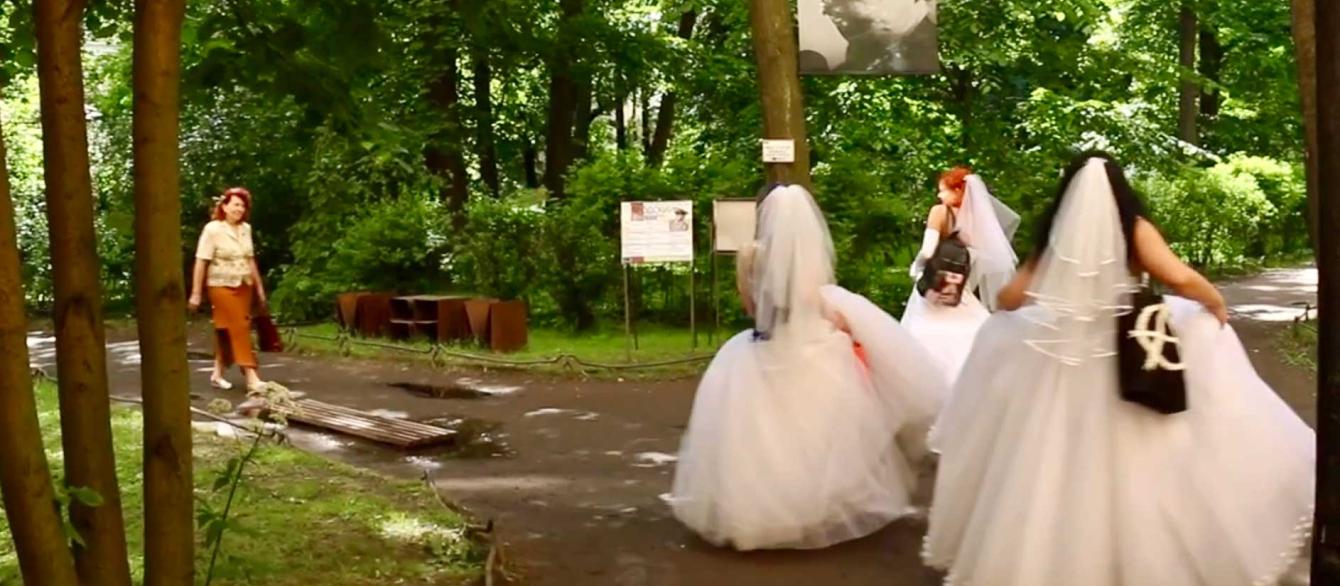 3 women in white wedding dresses with their backs to a viewer, the oethre woman walking towards them. Scene in the park, a lot of green trees