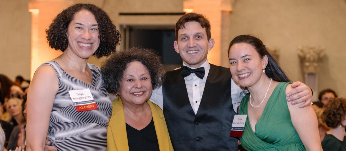 smiling instructor in yellow blazer flanked by three, also smiling, former students