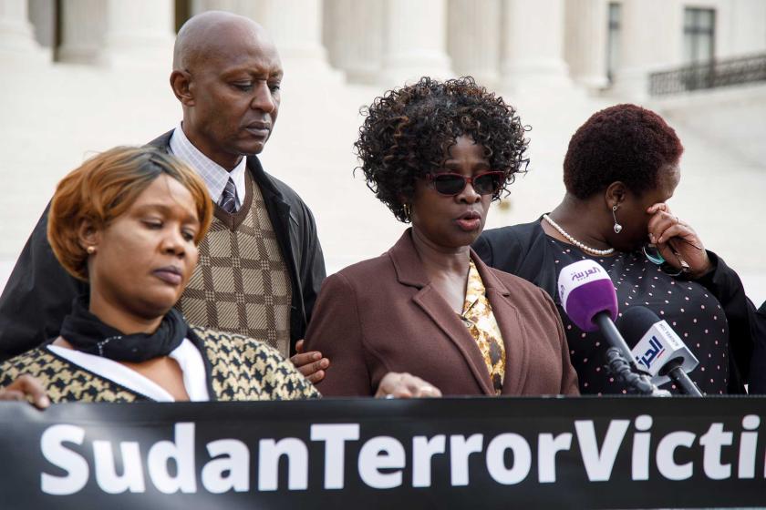 Three women and one man gather around a microphone at a news conference.  There is a banner in front of them reading "Sudan Terror Victims."