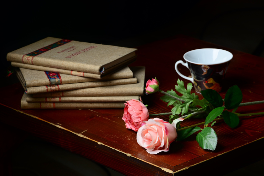 A stack of books next to a small mug and a few pink roses.