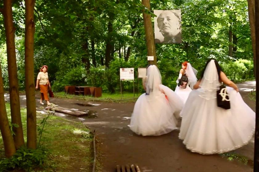 3 women in white wedding dresses with their backs to a viewer, the oethre woman walking towards them. Scene in the park, a lot of green trees